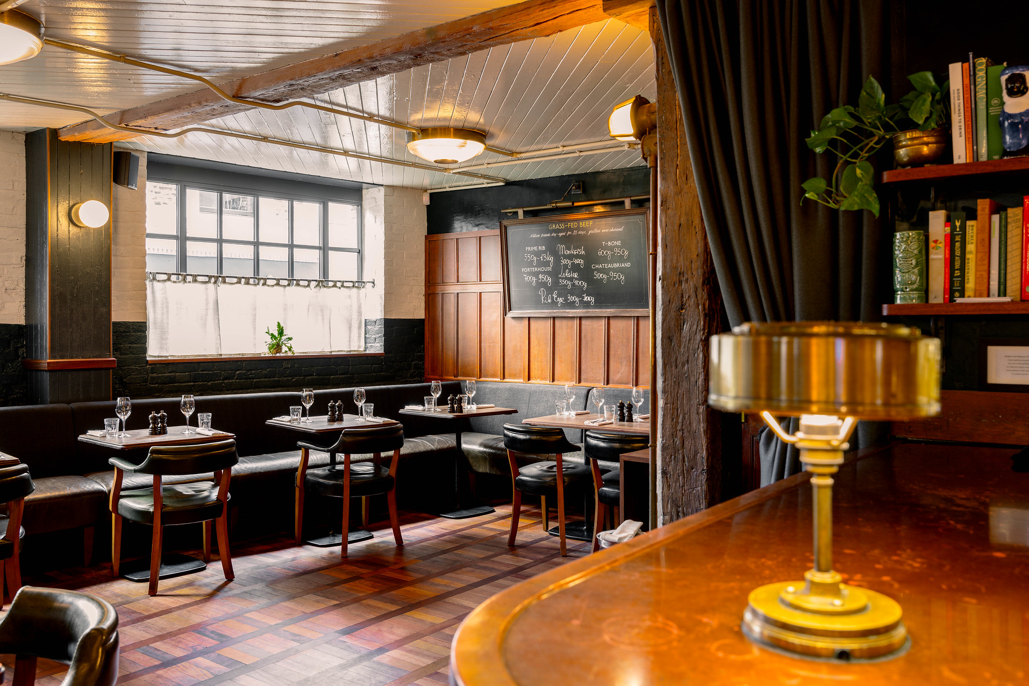Corner of Hawksmoor Borough with a brass lamp on a polished wood bar, bookshelves, a handwritten blackboard and banquette tables by the window