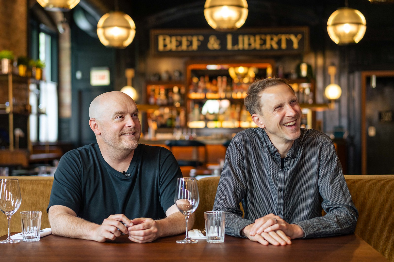 Founders Will and Huw sitting at a table, in Beef & Liberty Bar in Chicago, smiling, looking off to the left of the camera. They are dressed causally, one in a T-shirt and one in a button up shirt. On the table in front of each of them are two empty glasses.