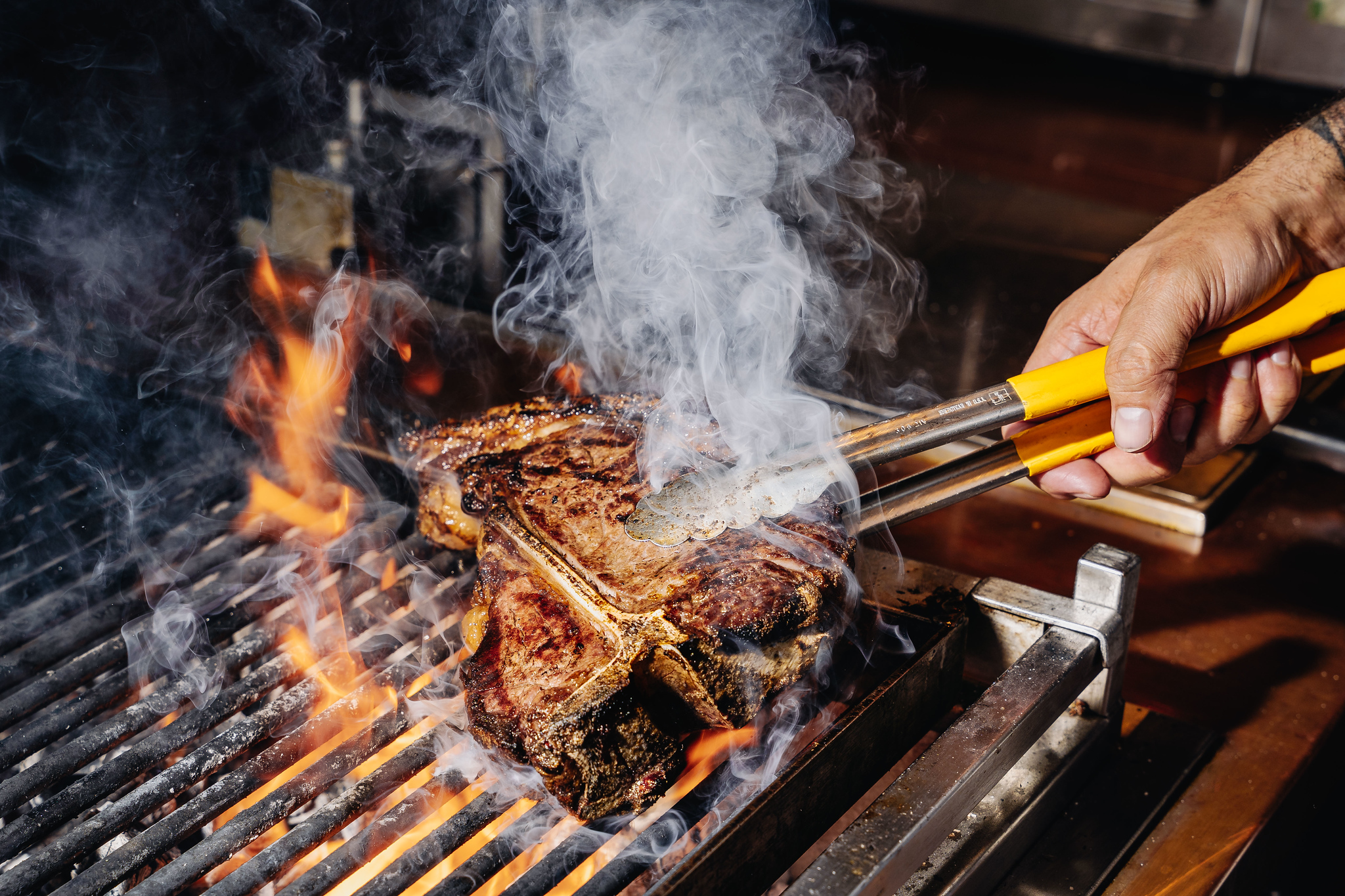 Chef turning a T-bone steak with tongs over flames on a charcoal grill
