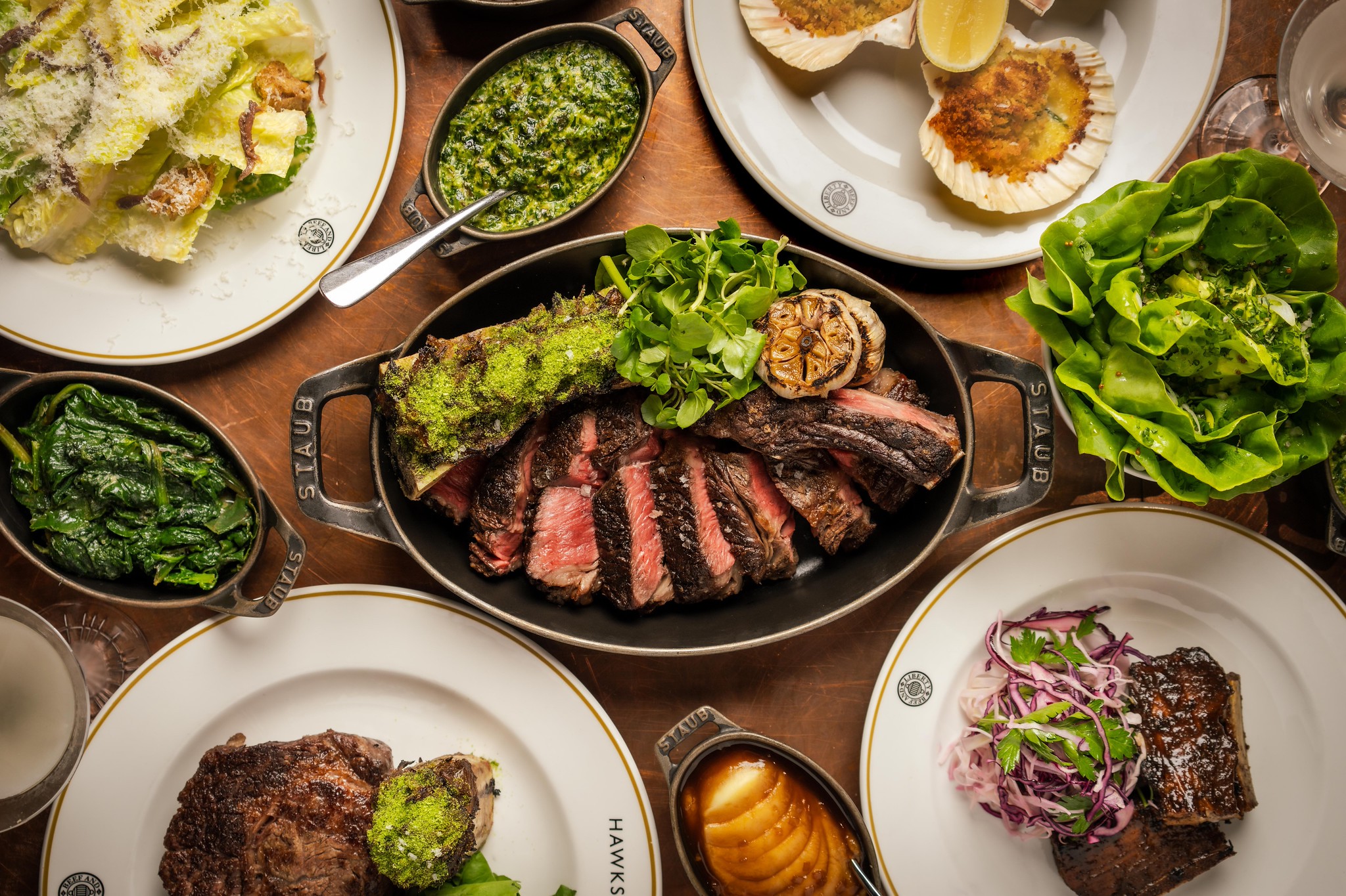 Overhead table of sharing dishes at Hawksmoor: sliced steak in a cast iron pan, scallops, greens, salads and sauces